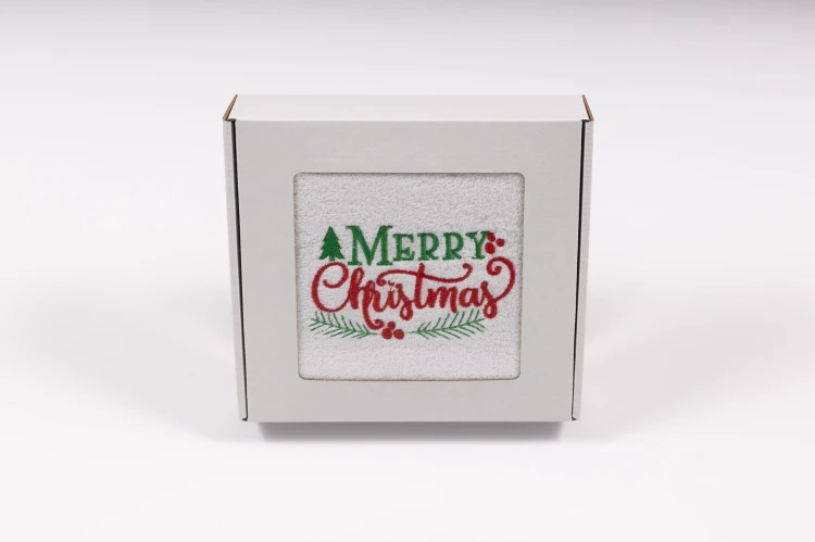 Elegancki świąteczny ręcznik frotte 500g z klasycznym haftem Merry Christmas i gałązkami ostrokrzewu, zapakowany w białe kartonowe pudełko z okienkiem, gotowy na prezent.