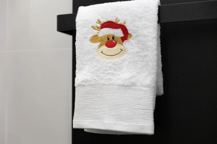 Miękki, bawełniany ręcznik bożonarodzeniowy z haftem wesołego renifera, powieszony na wieszaku w nastrojowej łazience jako świąteczna dekoracja wnętrza.