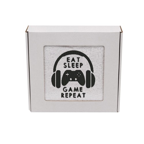 Gamingowy ręcznik z haftem pad, słuchawki i napis Eat Sleep Game Repeat zapakowany w ozdobne prezentowe pudełko z okienkiem.
