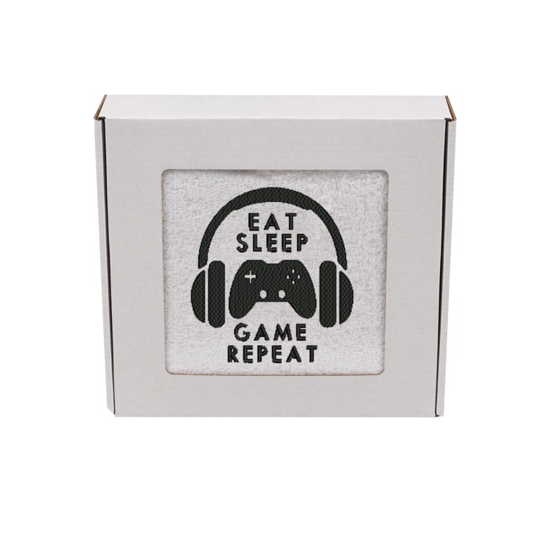 Gamingowy ręcznik z haftem pad, słuchawki i napis Eat Sleep Game Repeat zapakowany w ozdobne prezentowe pudełko z okienkiem.
