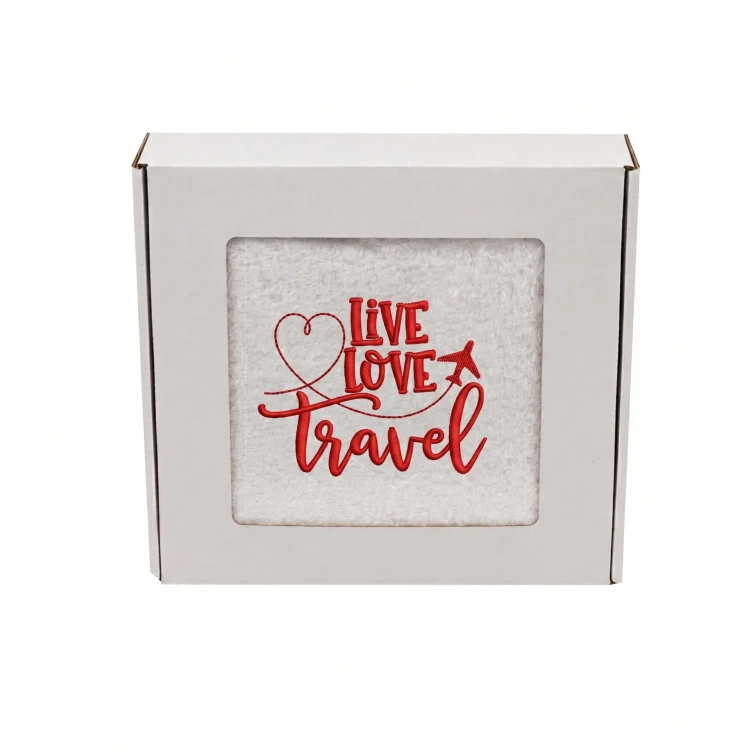 Biały ręcznik bawełniany z czerwonym haftem Live Love Travel oraz motywem samolotu i serca, zapakowany w ozdobne białe pudełko z okienkiem.