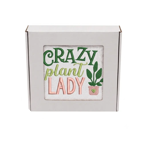 Biały bawełniany ręcznik frotte zapakowany w białe pudełko prezentowe z okienkiem. Przez okienko widoczny kolorowy haft: "CRAZY plant LADY" w kolorach zielonym, seledynowym i różowym oraz mała doniczka z sercem.