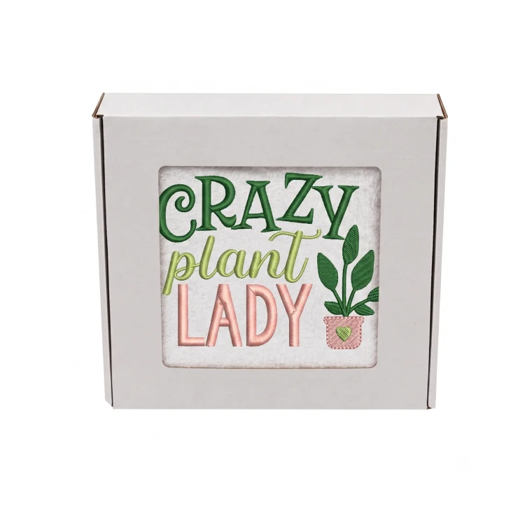Biały bawełniany ręcznik frotte zapakowany w białe pudełko prezentowe z okienkiem. Przez okienko widoczny kolorowy haft: "CRAZY plant LADY" w kolorach zielonym, seledynowym i różowym oraz mała doniczka z sercem.